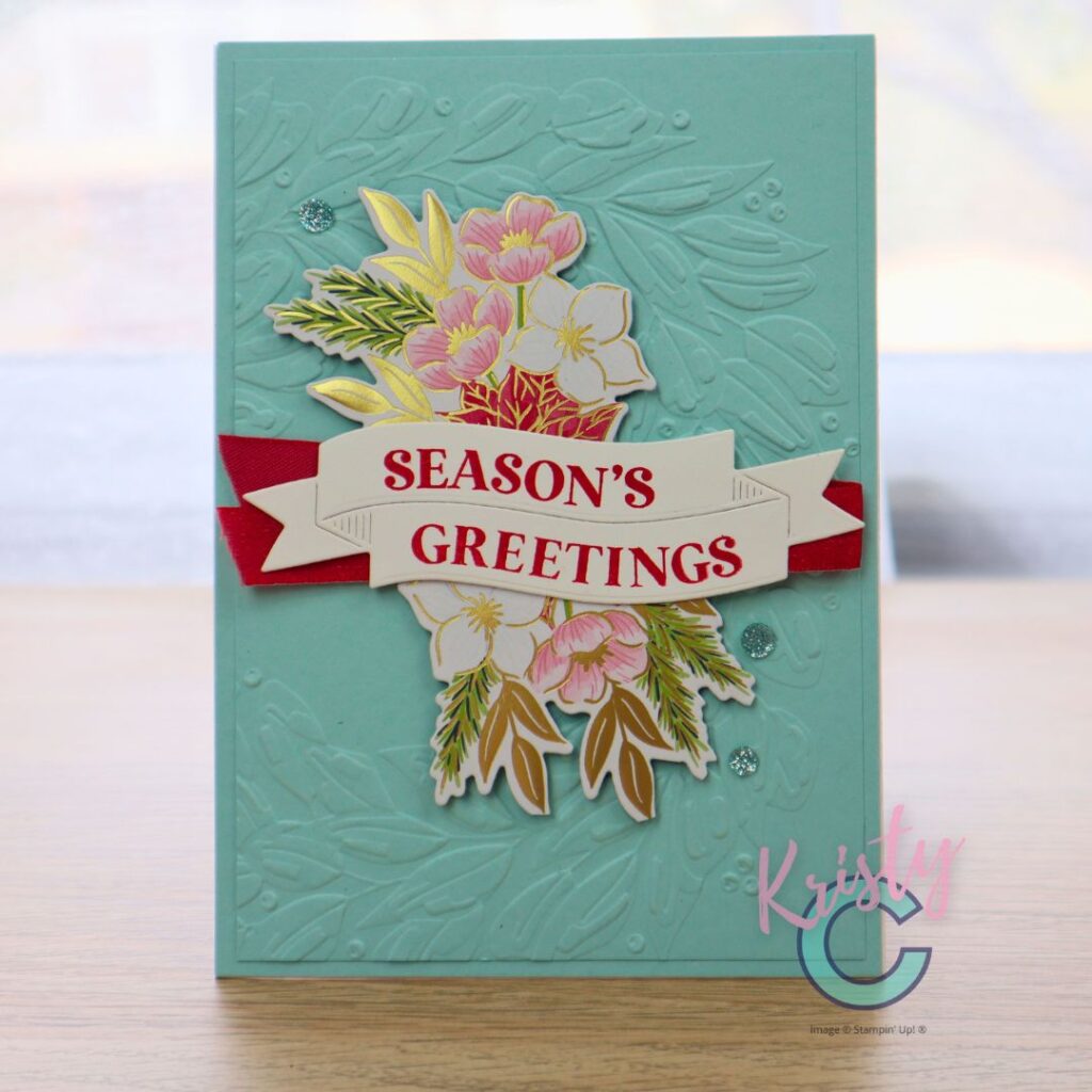 A handmade Christmas card featuring a turquoise dry-embossed background with a leafy texture, layered with red ribbon, gold-foiled floral elements, and a bold “Season’s Greetings” banner sentiment. Designed by Kristy Coromandel using Stampin’ Up! supplies.