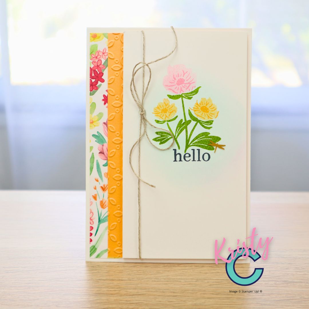 Handmade card with ink-blended background and stamped flowers in pink and yellow tones, accented with patterned paper strip and twine bow.