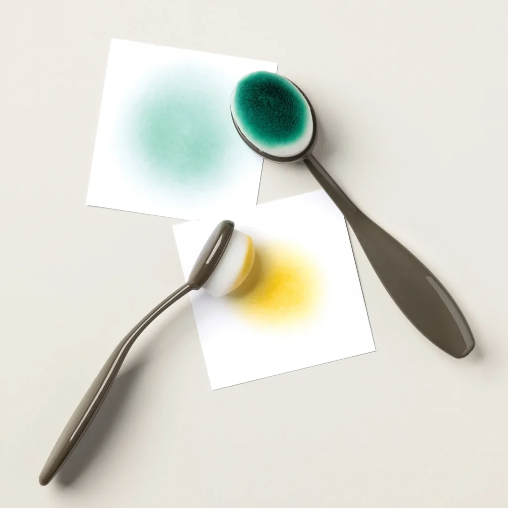 Two large blending brushes applying teal and yellow ink onto white cardstock squares in smooth circular motions.