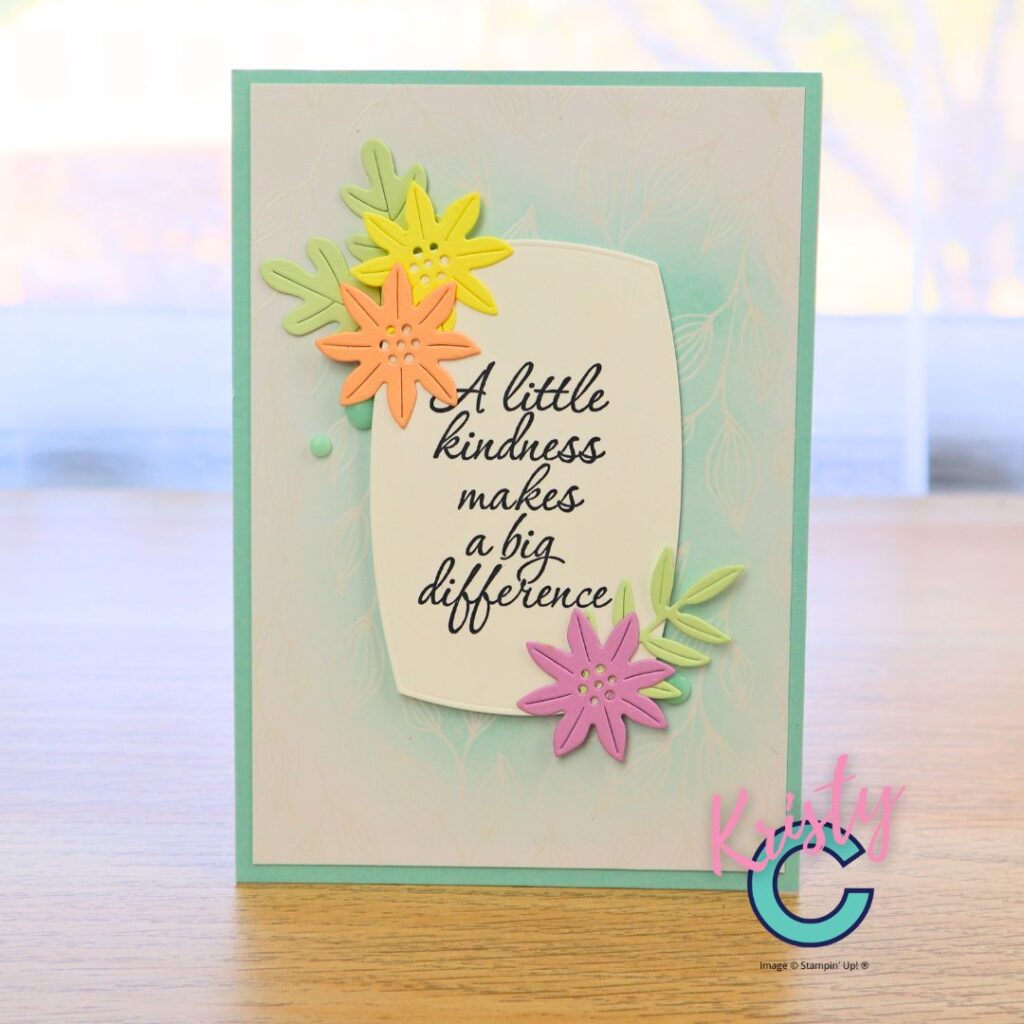 Handmade card with softly ink-blended background and pastel die-cut flowers surrounding a large sentiment panel reading “A little kindness makes a big difference.”