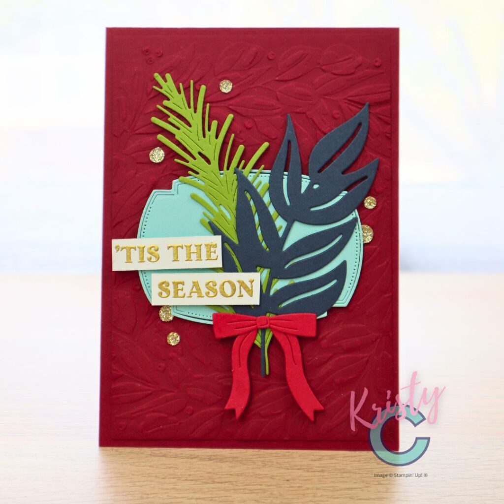 Red handmade holiday card featuring heat-embossed background with layered die-cut leaves, pine sprigs, and “’Tis the Season” sentiment in gold.