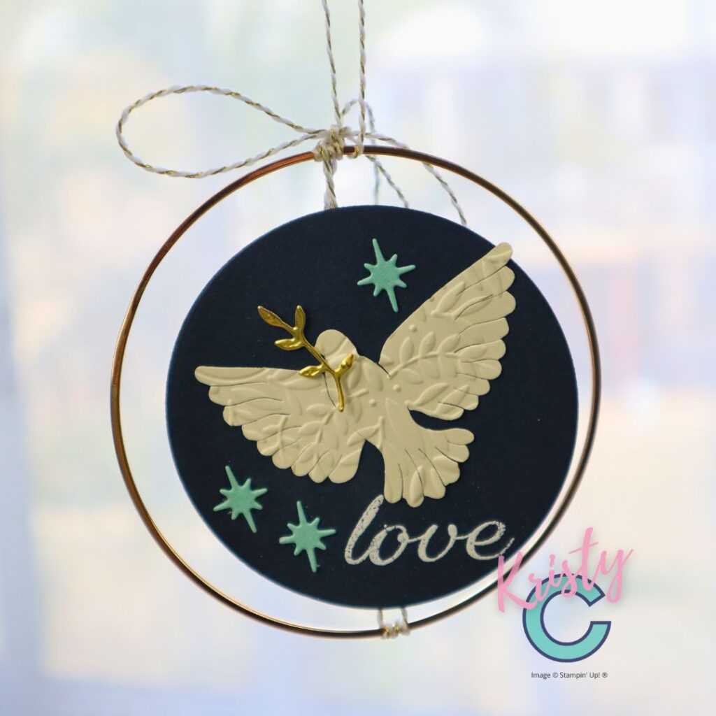 A handmade paper ornament featuring a cream embossed dove layered over a navy blue circular background, accented with mint stars, a gold olive branch, and the word “love” in silver script, hanging from a gold hoop with twine. Designed by Kristy Coromandel using Stampin’ Up! products.