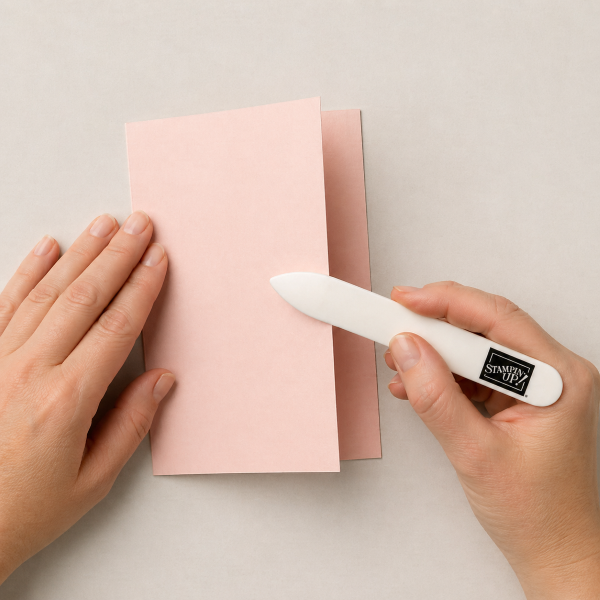 Hands using a bone folder to crease folded cardstock in half, creating a crisp fold for a standard greeting card.