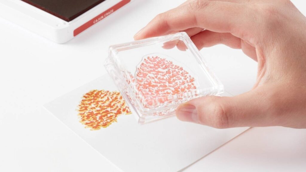 A hand holding a clear photopolymer stamp on an acrylic block above a stamped heart image on white cardstock, with a Cajun Craze ink pad in the background