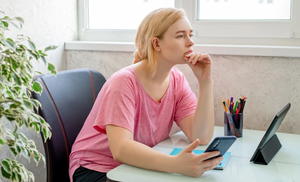 A woman in a pink top sitting at her desk, holding a phone and gazing out the window with a thoughtful expression — capturing the feeling of being stuck or unsure about your website.