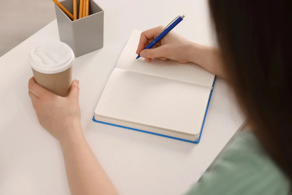 Close-up of a woman's hands writing in an open notebook while holding a takeaway coffee cup, suggesting the moment of getting clear and focused on what matters.