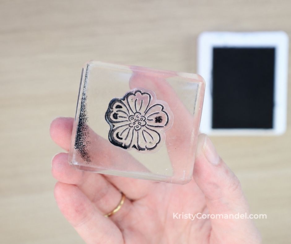 Overhead close-up of a clear photopolymer flower stamp mounted on an acrylic block, showing an even layer of ink across the stamped image before stamping onto cardstock.