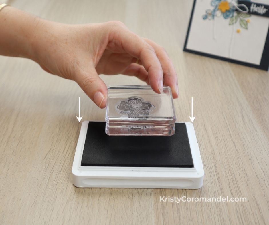 Hand holding a clear photopolymer stamp mounted on an acrylic block above an ink pad, demonstrating the correct stamp-to-ink technique for even ink coverage and cleaner stamped images.