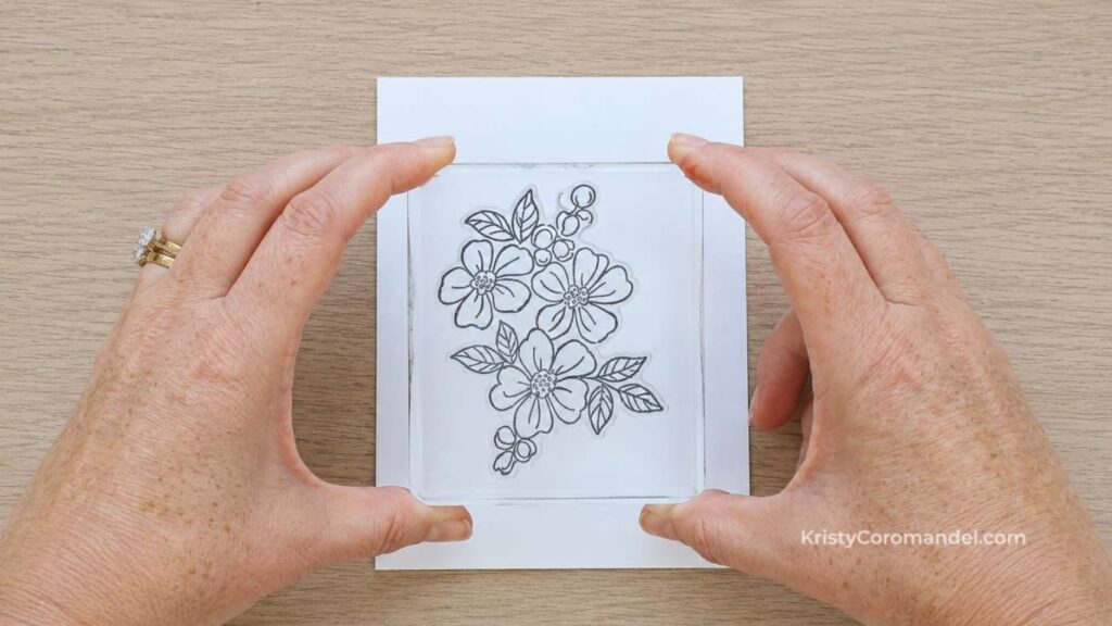 Overhead view of hands holding a clear acrylic block with a mounted photopolymer stamp above white cardstock, demonstrating how transparent stamps help with accurate positioning.