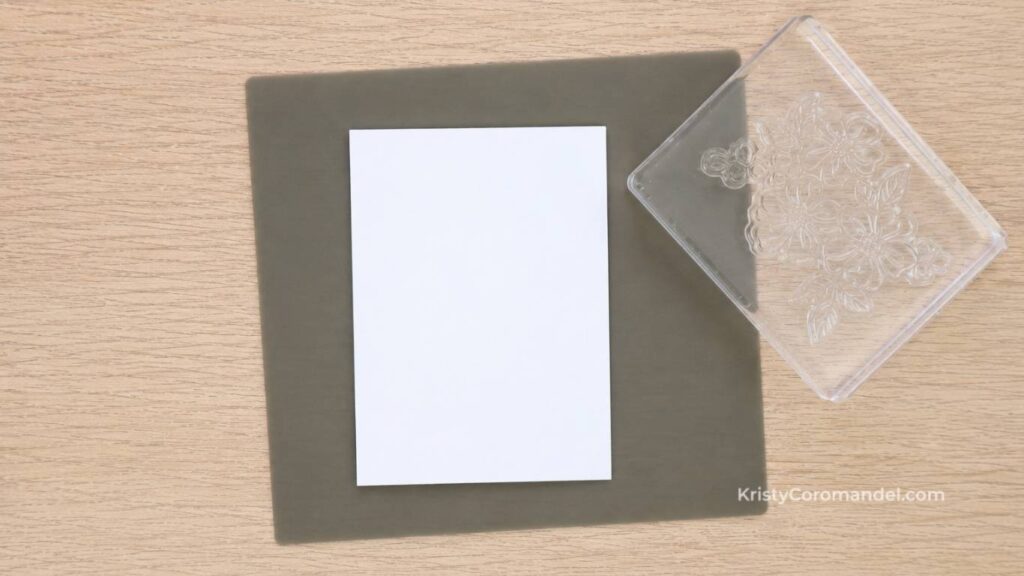 White cardstock placed on a silicone stamping mat beside a clear acrylic block with a mounted photopolymer stamp, ready for cardmaking.