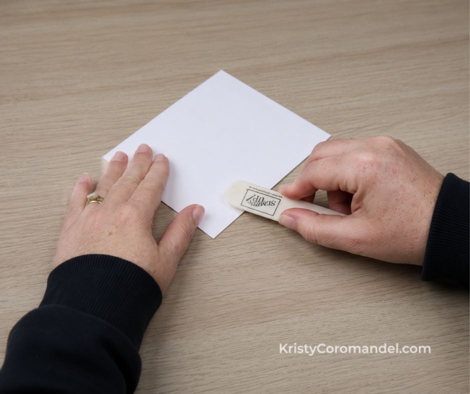 Hands using a Stampin' Up! bone folder to burnish the fold of a white folded cardstock card on a light wood crafting table.