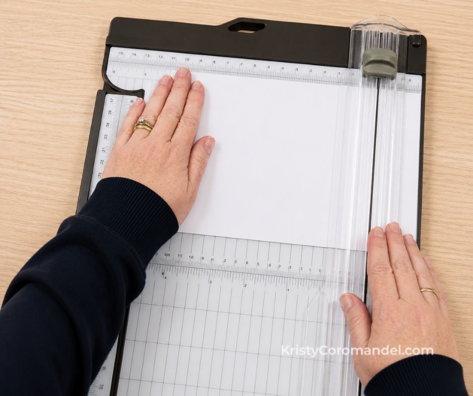 Hands positioning white cardstock against the top ruler of a paper trimmer, with one hand holding the cardstock firmly at the top while the other rests near the cutting blade to demonstrate correct hand placement.
