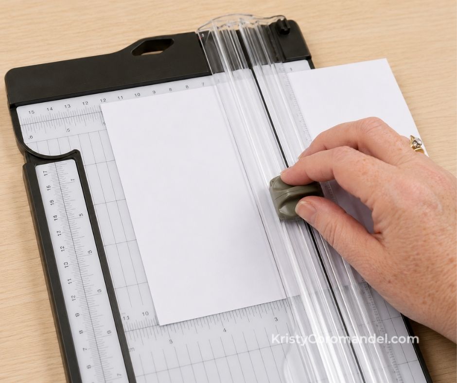 Close-up of a paper trimmer cutting white cardstock, with the cutting blade gliding smoothly down the track while a hand guides the slider for a clean, straight cut.