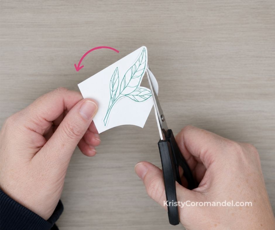 Top-down close-up of hands using detail scissors to fussy cut a stamped leaf image while rotating the cardstock for smoother, more controlled cuts.