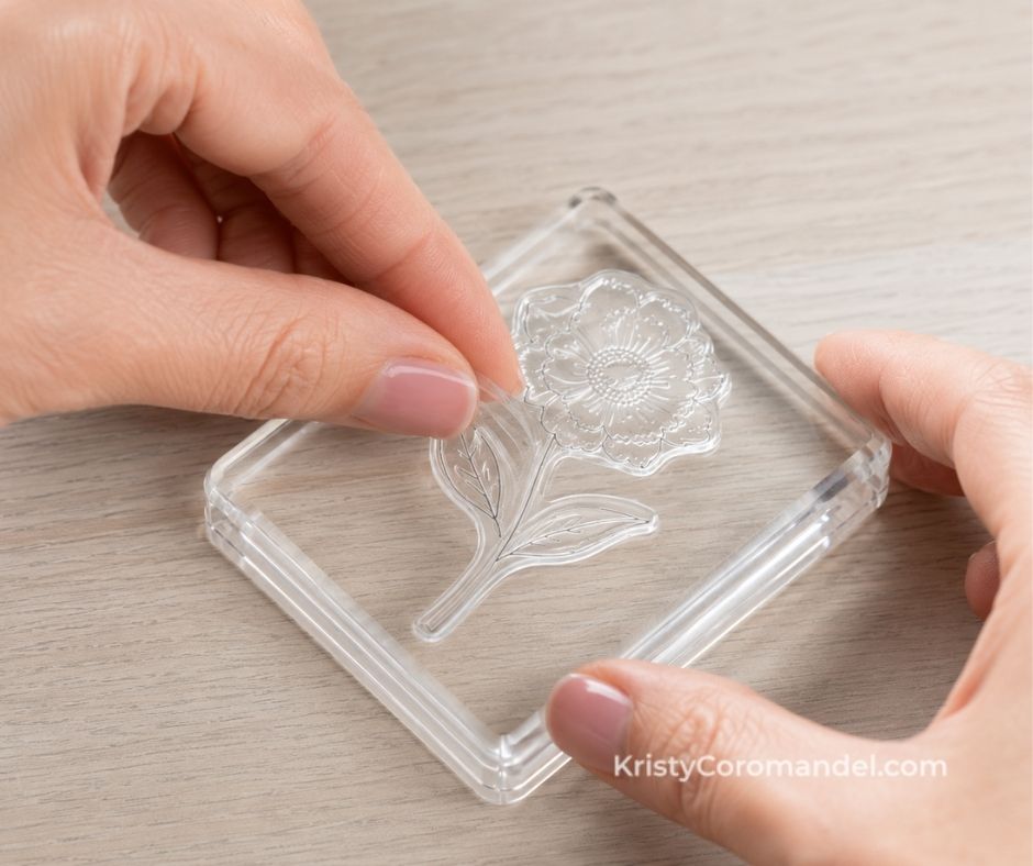 Close-up of hands peeling a clear photopolymer flower stamp onto a transparent acrylic stamping block on a light wood surface.
